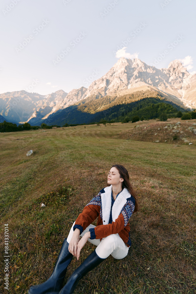 Naklejka premium woman sitting on meadow in mountain landscape, nature scene with relaxed pose and closed eyes, outdoors lifestyle with warm jacket and boots enjoying fresh air and peaceful view