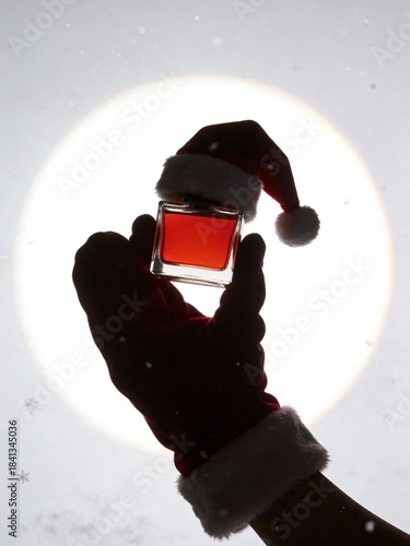 Silhouette of Hand in Santa Glove Holding Red Perfume Bottle Against Bright Circle