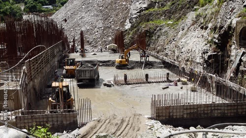 Yellow heavy equipment including excavators and lorries working at a dusty construction site in Nepal for a new hydropower station surrounded by metal reinforcement bars and walls.
