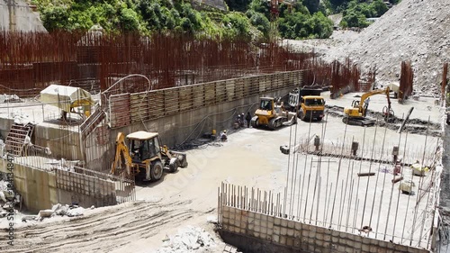 Active construction zone of a hydroelectric dam in Nepal displaying heavy earthmoving machinery and complex steel rebar framework for a massive concrete infrastructure foundation.