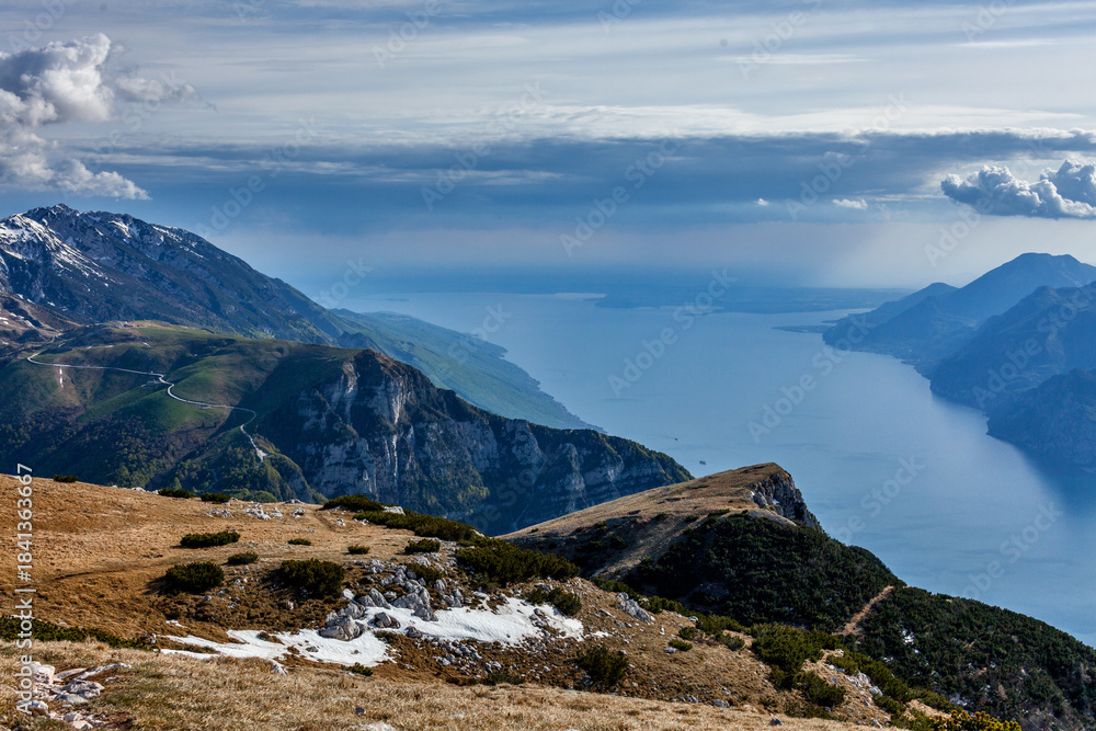 Ταπετσαρία Wide view of Lake Garda from Monte Altissimo—alpine terrain, winding roads, and distant water under a partly cloudy sky