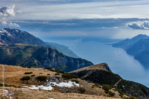 Canvas Print Wide view of Lake Garda from Monte Altissimo—alpine terrain, winding roads, and distant water under a partly cloudy sky