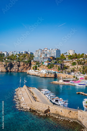A vibrant high-resolution image of Antalya’s scenic marina, featuring docked boats, a stone pier, turquoise Mediterranean waters, and dramatic coastal cliffs lined with modern buildings