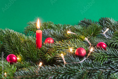 Christmas burning red candle in between fir branches with Christmas tree balls and little lights on green