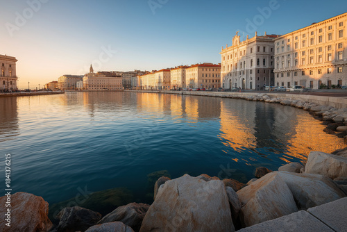 Sunlit waterfront of Trieste Italy with historic architecture reflected in calm harbor water at golden hour