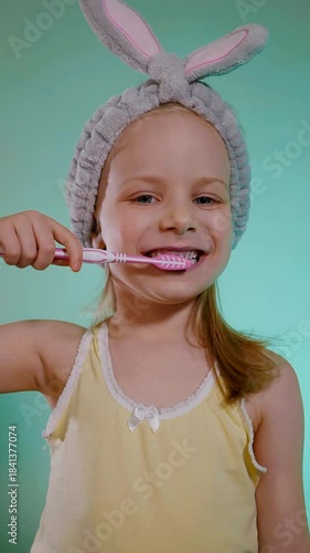 Child with rosy cheeks and a yellow tank top vigorously brushing, showcasing morning routine fun. Close-up portrait of a smiling child using a pink and white toothbrush, fuzzy bunny ears poised above 