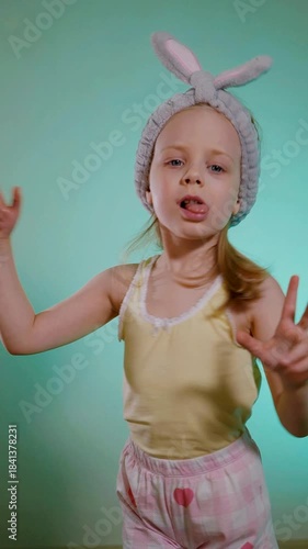 Vertical. Young child with bunny ears headband singing and dancing funny against a bright cyan background. Toddler in yellow tank top and pink heart-patterned shorts