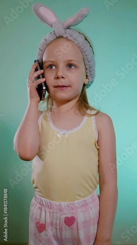 Vertical. Young girl with bright blue eyes wearing a fluffy gray bunny-ear headband talks on a black smartphone against a teal background. Child in yellow tank top and pink heart-patterned pajama 
