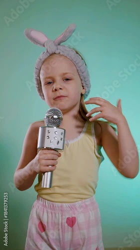 Vertical. Young child with bunny ears headband singing into a sparkling silver microphone against a bright cyan background. Toddler in yellow tank top and pink heart-patterned shorts holding a karaoke