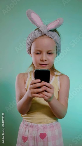 Vertical. Young girl with a fluffy gray bunny ear headband smiling while looking intently at a black smartphone against a bright turquoise background.