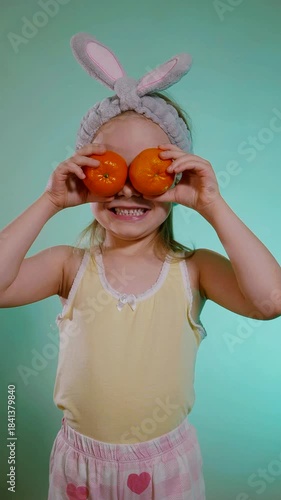Vertical. Cheerful young girl wearing a fuzzy gray bunny ear headband holds two bright orange tangerines over her eyes, grinning widely against a vibrant teal background.