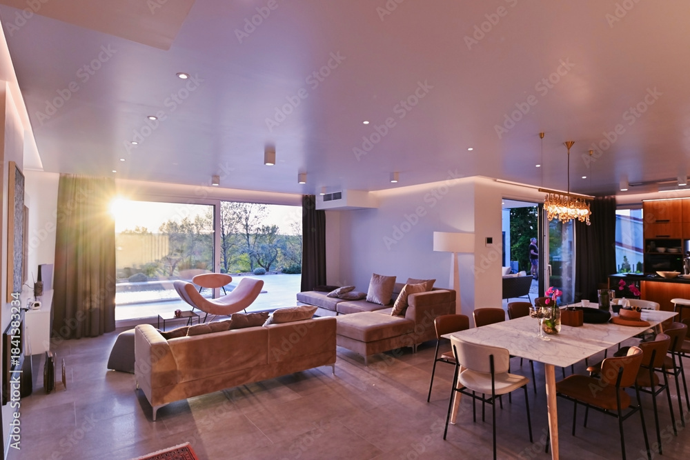 Obraz premium Modern luxury living room interior at sunset. Open-plan space featuring beige sofas, marble dining table, large glass windows with sun flare, and view of a patio. Stylish contemporary home design