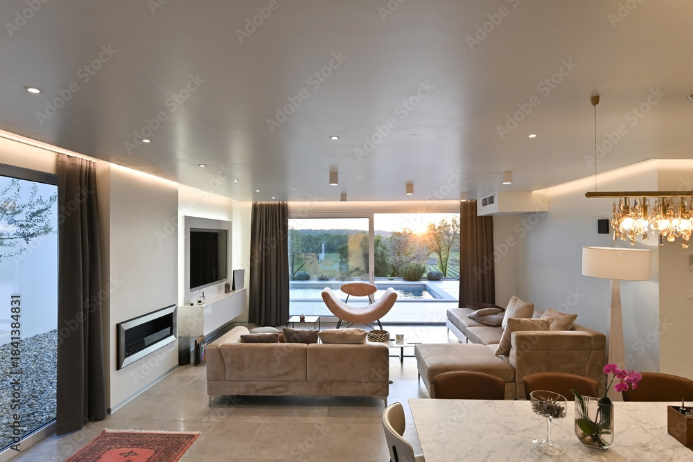 Obraz premium Modern luxury living room interior at sunset. Open-plan space featuring beige sofas, marble dining table, large glass windows with sun flare, and view of a patio. Stylish contemporary home design