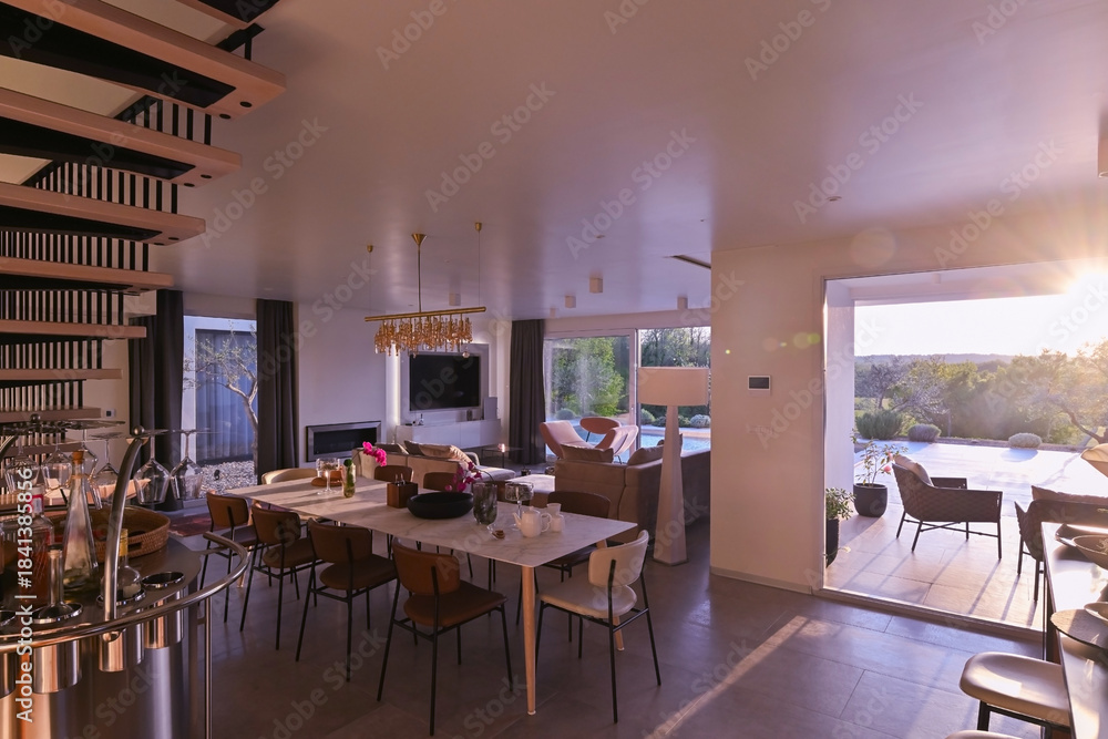 Obraz premium Modern luxury living room interior at sunset. Open-plan space featuring beige sofas, marble dining table, large glass windows with sun flare, and view of a patio. Stylish contemporary home design