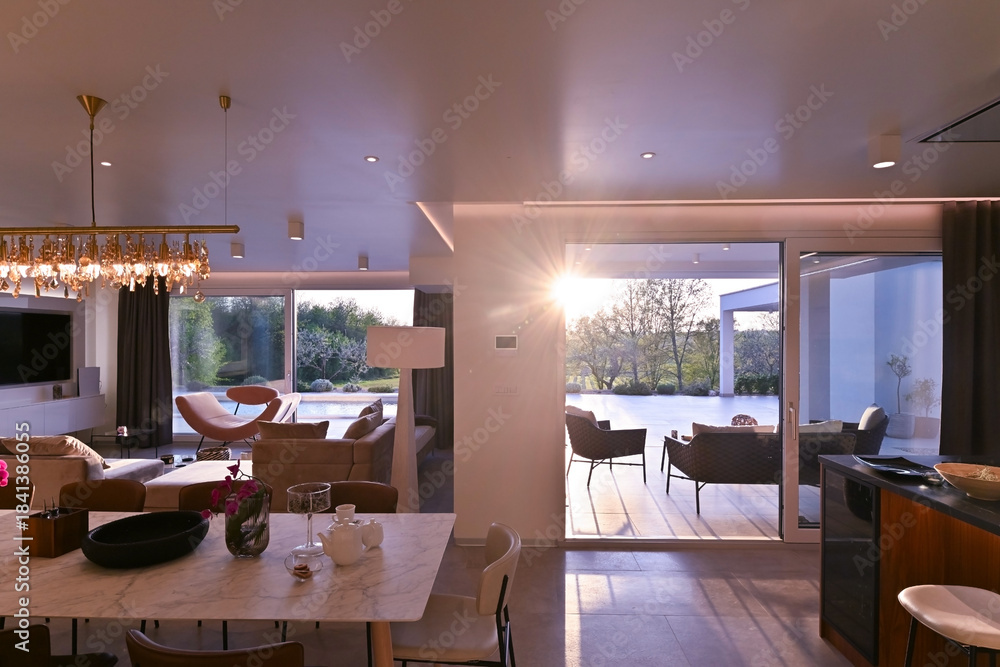 Obraz premium Modern luxury living room interior at sunset. Open-plan space featuring beige sofas, marble dining table, large glass windows with sun flare, and view of a patio. Stylish contemporary home design