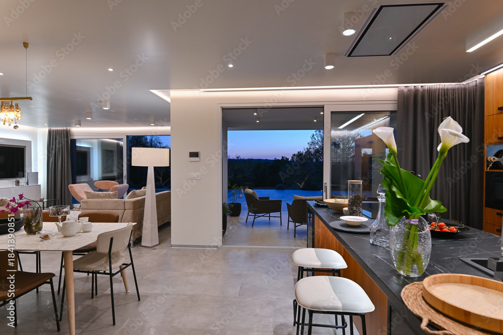 Obraz premium Modern luxury living room interior at sunset. Open-plan space featuring beige sofas, marble dining table, large glass windows with sun flare, and view of a patio. Stylish contemporary home design