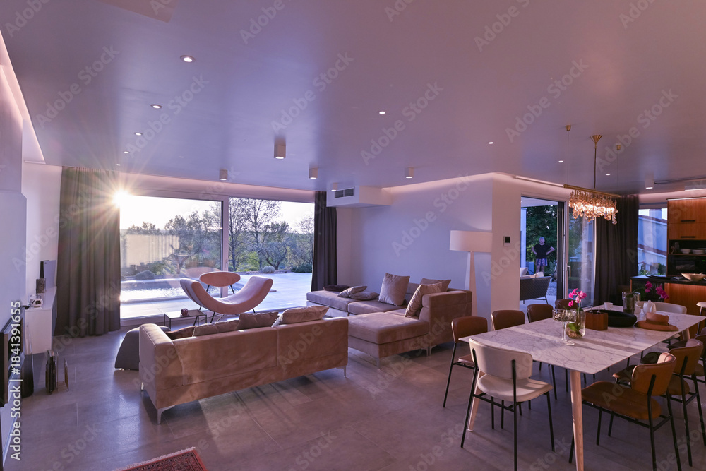 Obraz premium Modern luxury living room interior at sunset. Open-plan space featuring beige sofas, marble dining table, large glass windows with sun flare, and view of a patio. Stylish contemporary home design