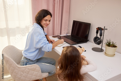 Mother working on a laptop while daughter plays with a teddy bear at home