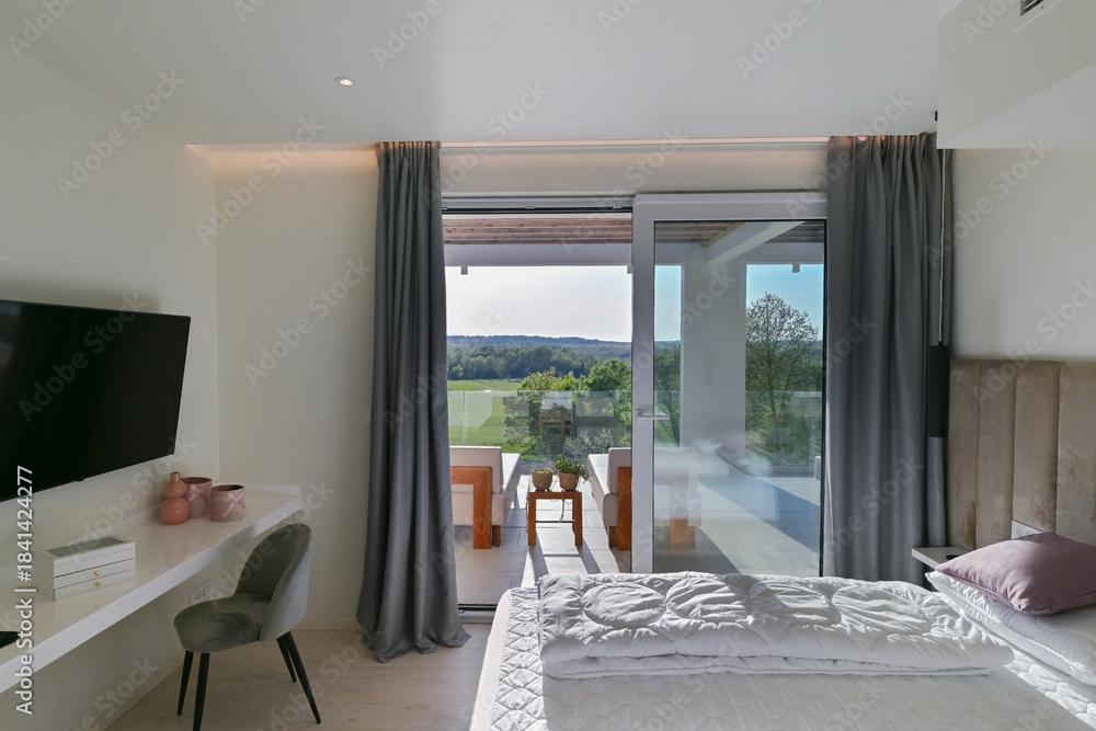 Naklejka premium Bright, modern bedroom featuring a comfortable bed, sleek desk, and a large sliding glass door opening to a private balcony with stunning green landscape views, perfect for relaxation and serene