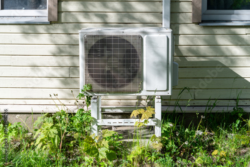 External heat pump unit positioned on ground beside wall of old wooden house, forming contrast between modern equipment and aged structure