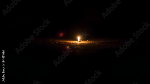 Single small candle burning brightly in the absolute darkness, creating a warm glow on the ground