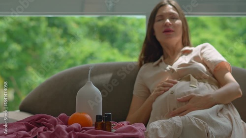 Pregnant woman over 40 sitting on a couch by a sunlit window enjoying scent and relaxation from a working electric aroma diffuser. Wellness, pregnancy care and relaxation lifestyle concept. Slowmotion