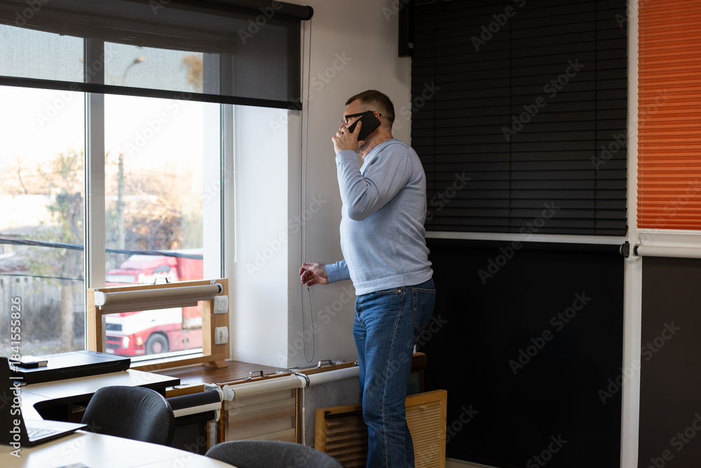 Fototapeta premium Small business owner of a blinds retail shop standing by a window with a roller shade, talking on the phone while handling work tasks. Modern interior, natural light and professional atmosphere.