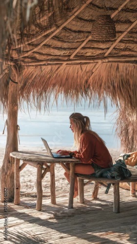 Woman working on laptop at the beach under a thatched roof structure