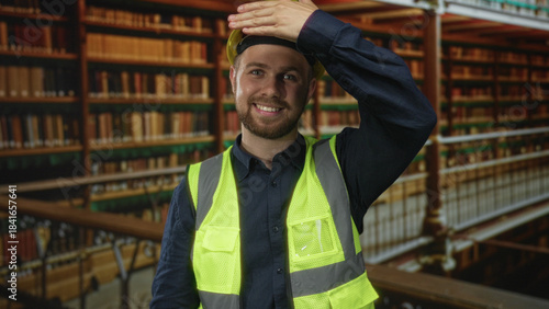 Vászonkép Man builder in yellow hardhat and neon high vis vest covers mouth with hand among books on shelves in library building, eyes closed; silence study focus