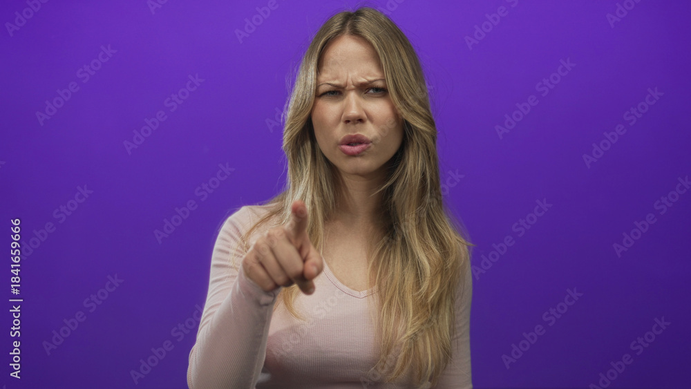 Fototapeta premium Blonde caucasian woman points finger at camera in purple studio with serious expression; accusation anger.