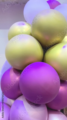 A festive cluster of colorful party balloons in shades of metallic gold, matte white, and vibrant purple. The balloons are arranged in a dense bunch, showing smooth textures and soft light reflections