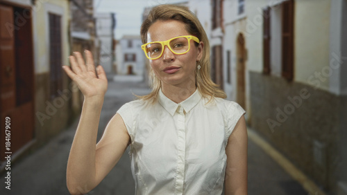 Fotografie Woman wearing white shirt and yellow glasses waves hand in sunlit old town street framed by stone buildings; welcome