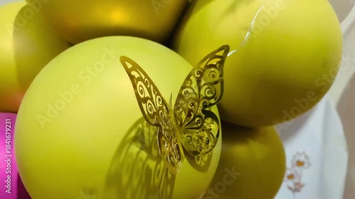 A close-up, warm-toned photograph of a delicate, filigree-style decorative butterfly. The butterfly is made of gold or brass-toned metal, featuring intricate, laser-cut swirl and scroll patterns on it