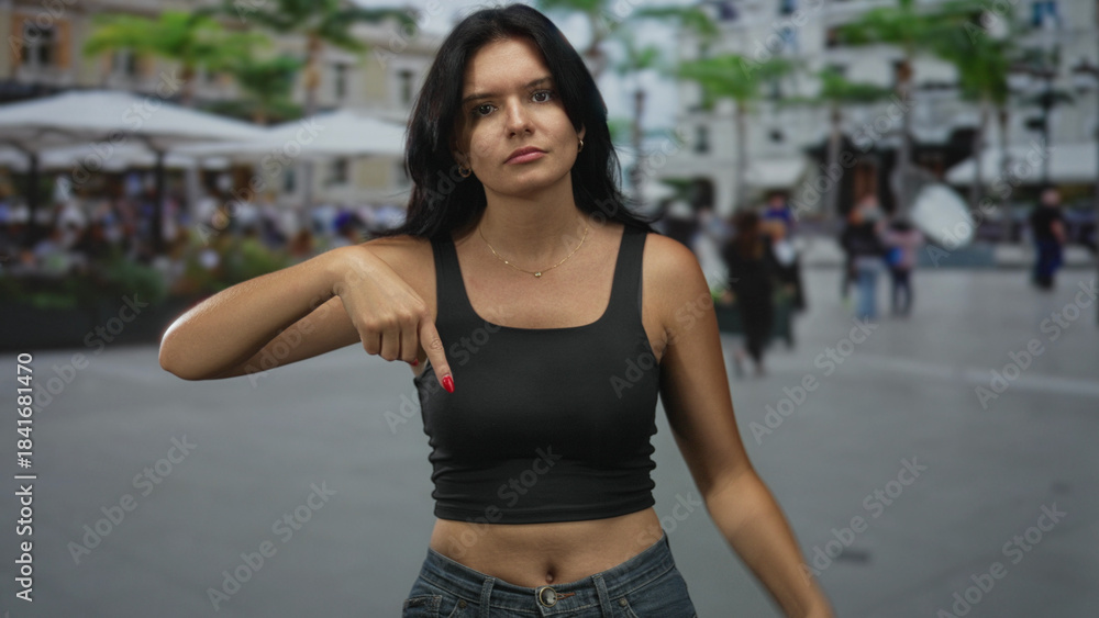 Fototapeta premium Young hispanic woman pointing finger to navel and exposed midriff on busy street; body positivity confidence.