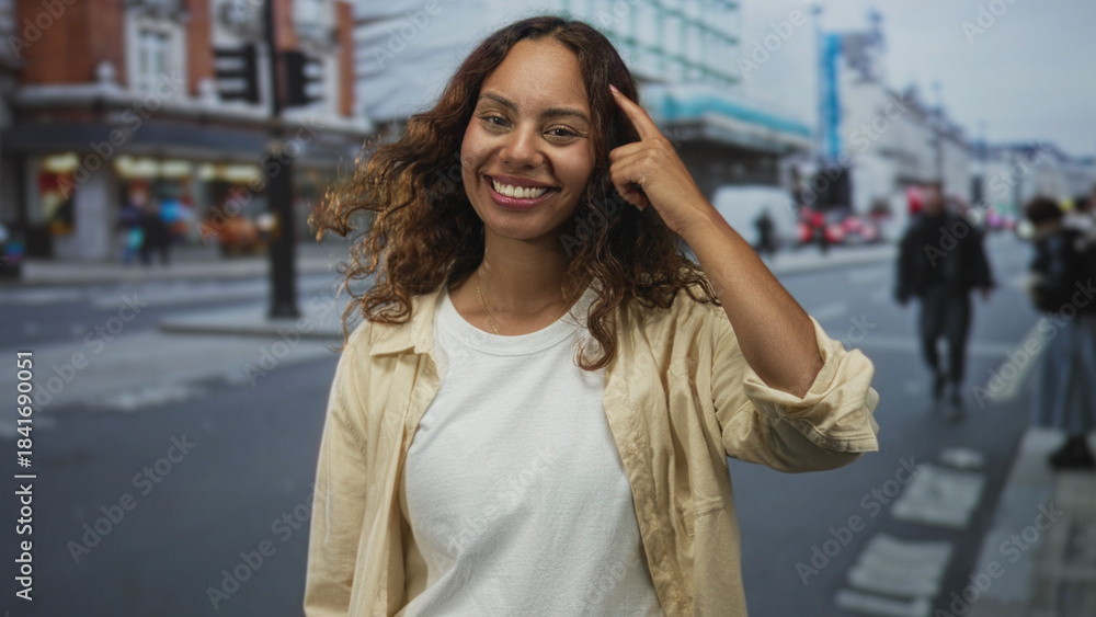 Fototapeta premium Woman points finger to temple on urban city street, smiling at camera while wearing white t shirt and beige overshirt, curly hair framing face; confidence.