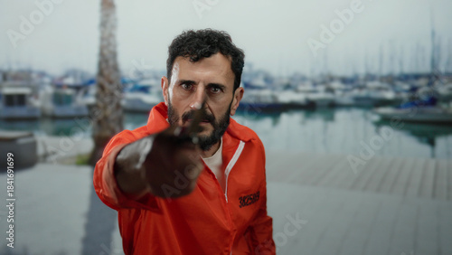 Fotografi Hispanic man with beard in prison jumpsuit holding knife at seaside promenade with boats and palm tree visible