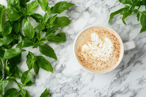 Top view of creamy latte in white cup on marble surface surrounded by fresh leaves. Rich coffee foam and vibrant green herbs, bright, appetizing culinary composition with natural, organic atmosphere