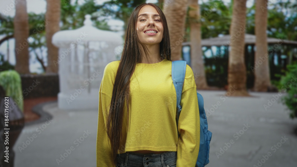 Fototapeta premium Teenage girl in yellow sweater with blue backpack standing with head tilt and subtle smile in park; youthful optimism.