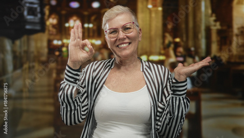 Obraz na plátně Middle aged woman smiling, showing ok sign with raised hands inside a church building interior, wearing glasses and striped shirt; joy gratitude