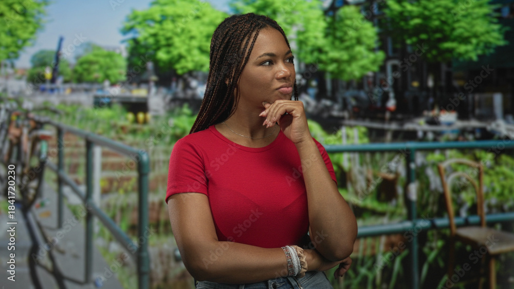 Fototapeta premium Woman with hand on chin on street canal railing in green park wearing red tshirt and bracelet; thoughtful contemplation.