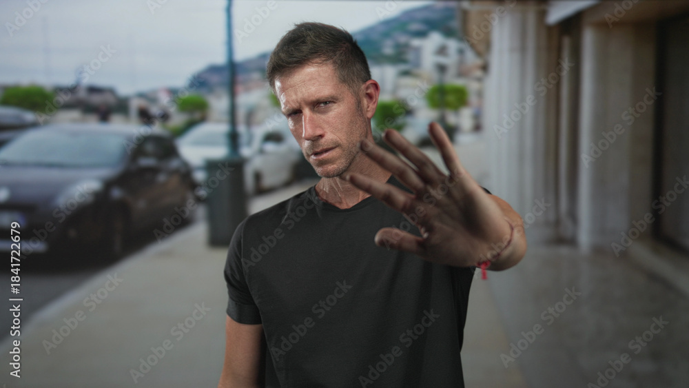 Fototapeta premium Man with hands raised palms out on street blocking forward movement, facing camera; caution pause restraint.