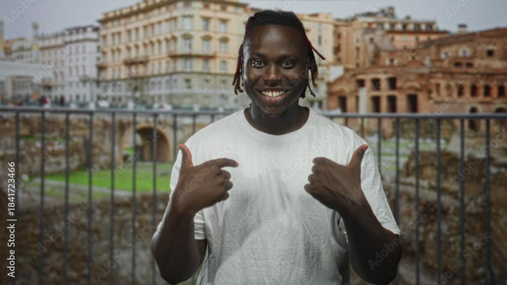Φωτογραφία Man with thumbs pointing to chest at historic roman ruins building, smiling confidently to camera; pride travel memory