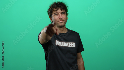 Фотография Hispanic man in volunteer shirt pointing forward against green background, conveying enthusiasm and determination for service and helping others with a confident gesture
