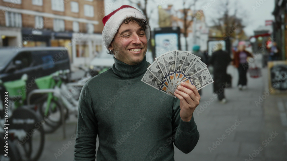 Fototapeta premium Man wearing christmas hat smiling while holding dollars on street outdoors during holiday season with blurred background and people walking by.