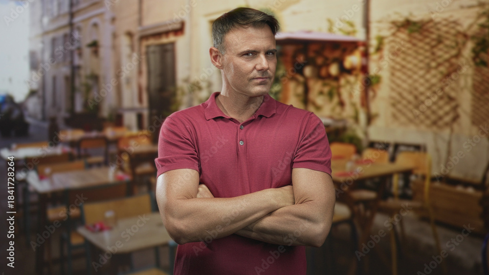Obraz premium Man with arms crossed showing forearms, wearing a red polo, standing in a street cafe near tables and a storefront; confidence calm resolve.