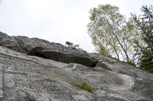 Rugged rock wall of Written Stone rises beside pine and deciduous tree line featuring faint graffiti. Autumn colors tint the scene conveying solitude as natural and urban marks meet on remote hillside