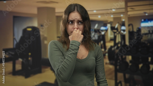 Billede på lærred Woman pinching nose with hand and looking worried while standing in gym building near dumbbells and cardio machines; disgust unease