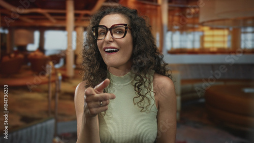 Fotografie Woman pointing finger in hotel lounge with open palms and bright smile against plush couches and ambient lighting; hospitality