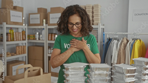 Photos Middle-aged hispanic woman volunteer in green uniform working in charity indoor room surrounded by donation boxes and meal trays, showing appreciation