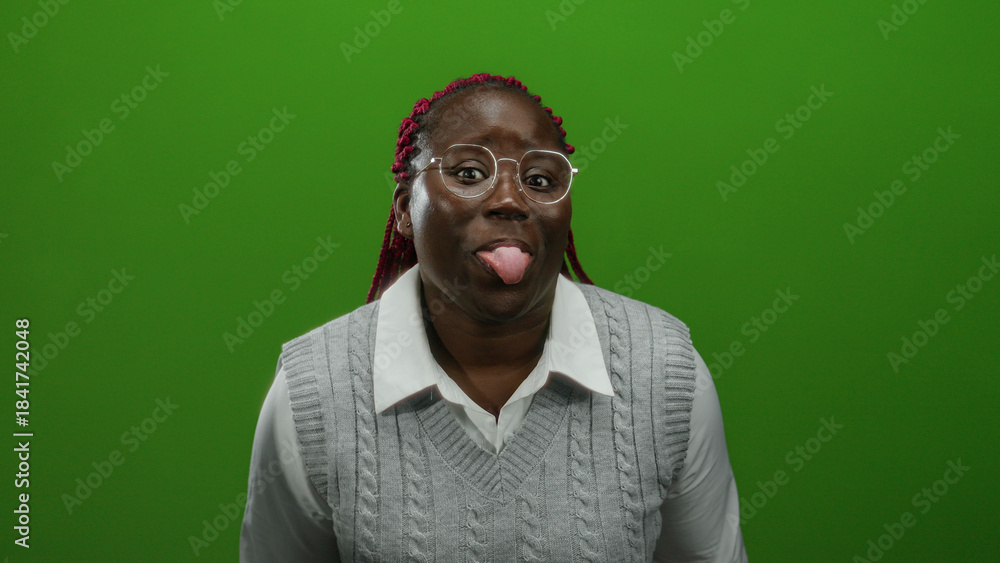 Fototapeta premium Woman with glasses and braided hair playfully sticking tongue out against a vibrant green wall background, wearing a gray sweater and white shirt.